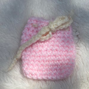 May include: A small, rectangular, light pink crocheted pouch featuring a textured stitch pattern. The pouch is secured at the top with a cream-colored crocheted drawstring cord that is tied into a simple knot across the front surface.