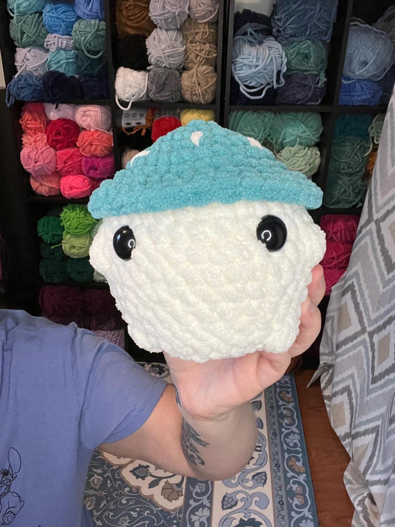 May include: A handmade, plush mushroom toy with a white body and a light blue cap. The cap has white polka dots and the mushroom has large, black button eyes. The toy is made of soft, textured yarn.