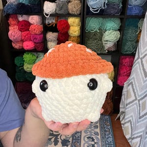 May include: A handmade, plush mushroom toy with an orange cap and white spots, and a white body. The mushroom has large, black button eyes. The background shows shelves filled with colorful yarn skeins, suggesting a craft or hobby theme.