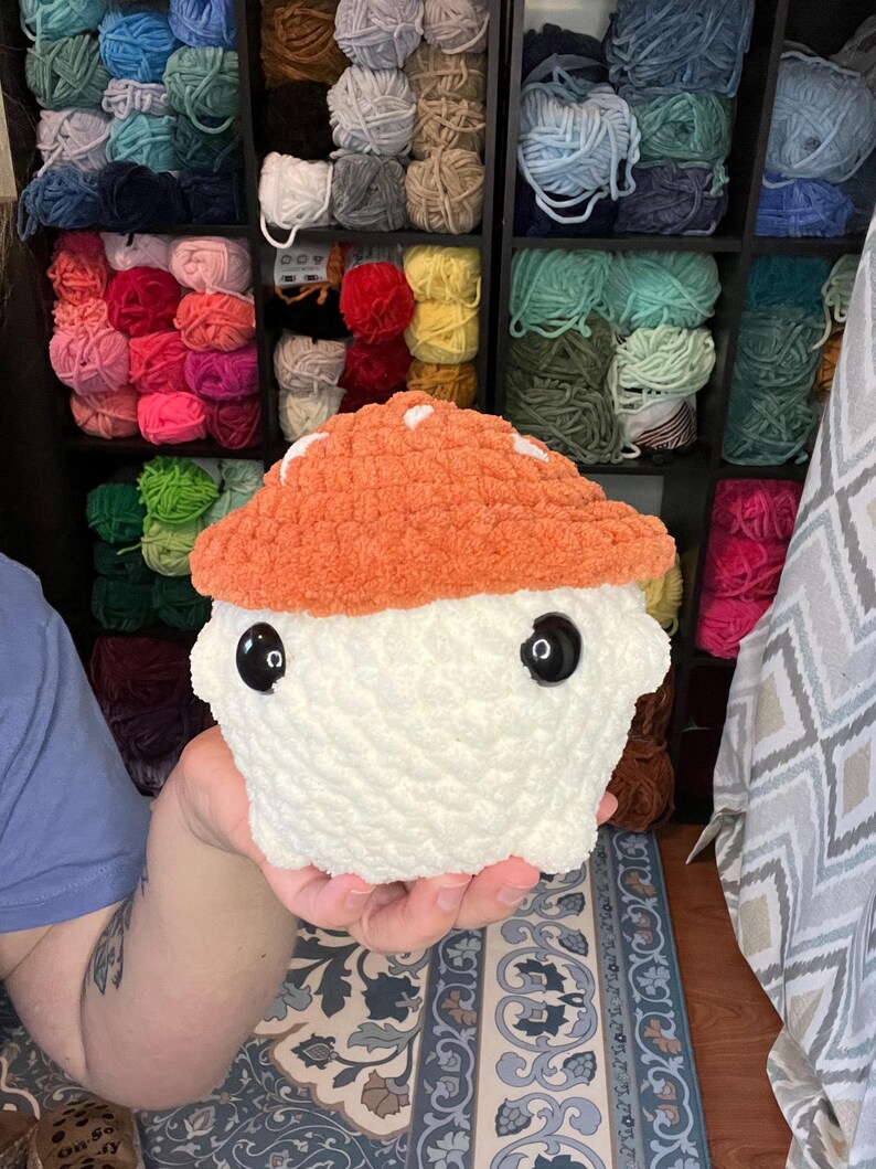 May include: A handmade, plush mushroom toy with an orange cap and white spots, and a white body. The mushroom has large, black button eyes. The background shows shelves filled with colorful yarn skeins, suggesting a craft or hobby theme.