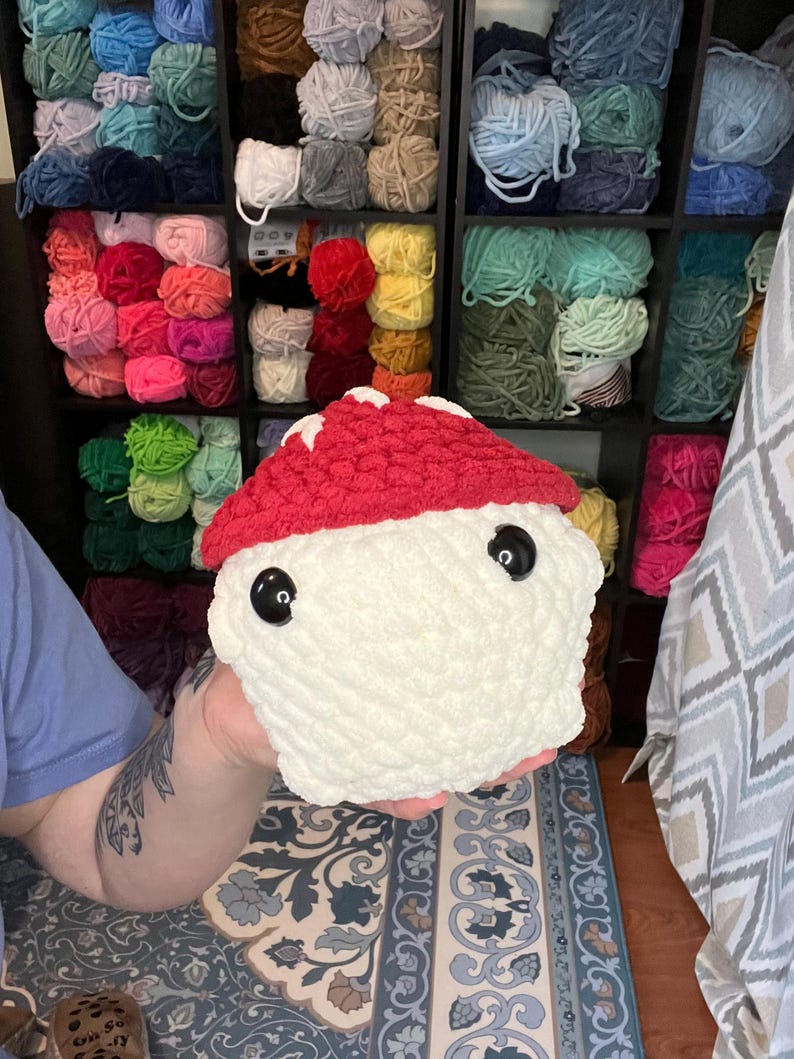 May include: A handmade, plush mushroom toy with a red cap and white stem. The mushroom has large, black button eyes and is made of soft, textured yarn. The background shows shelves filled with colorful yarn skeins.