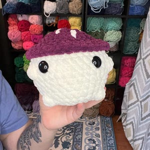 May include: A handmade, plush mushroom toy with a white body and a deep purple cap. The cap features small white spots. The mushroom has large, black, button eyes. The background includes shelves filled with colorful yarn.