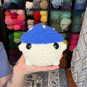 May include: A handmade, plush mushroom toy with a blue cap and white body. The mushroom has large, black button eyes and white star accents on the cap. The background shows shelves filled with various colors of yarn.