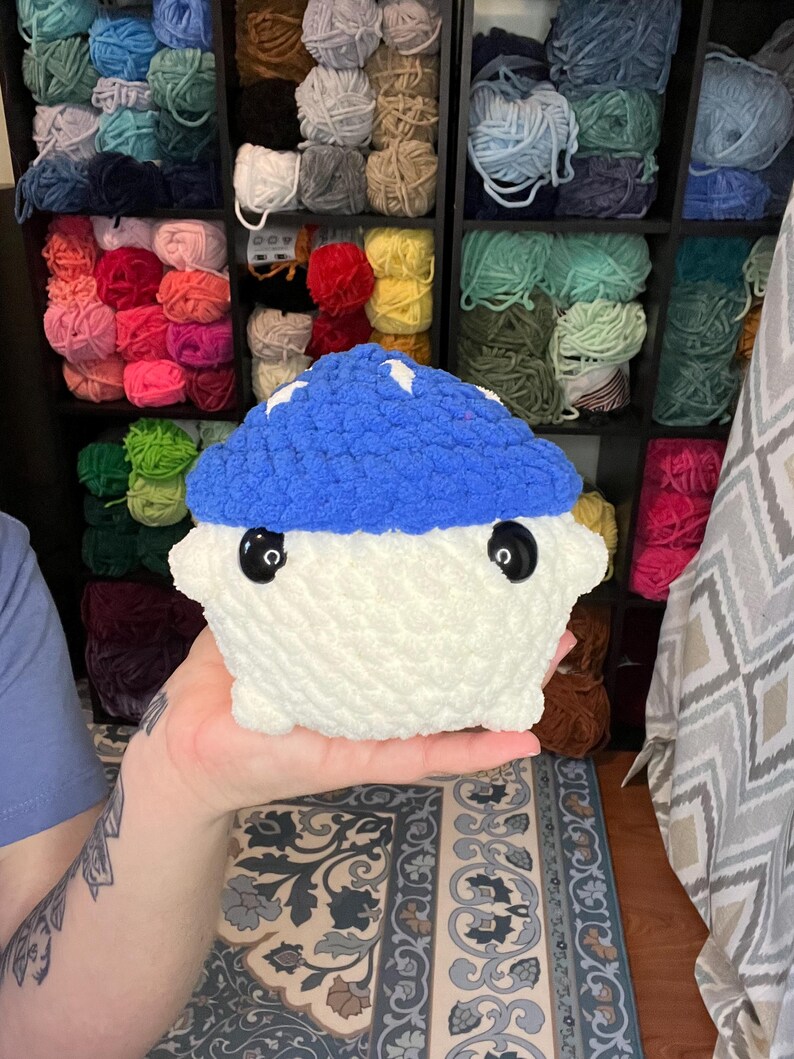 May include: A handmade, plush mushroom toy with a blue cap and white body. The mushroom has large, black button eyes and white star accents on the cap. The background shows shelves filled with various colors of yarn.