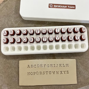 May include: A white plastic case with a brown label that says "DirtKicker Tools" contains a set of 26 brown rubber stamps with white letters of the alphabet. The stamps are arranged in a white plastic tray. A piece of brown leather is below the tray with the alphabet stamped on it.