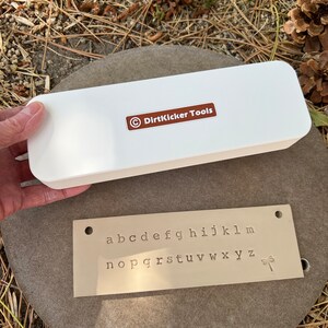 May include: A white rectangular box with the text "DirtKicker Tools" and a metal plate with stamped lowercase letters a-z and a dragonfly symbol. The items are on a gray stone.