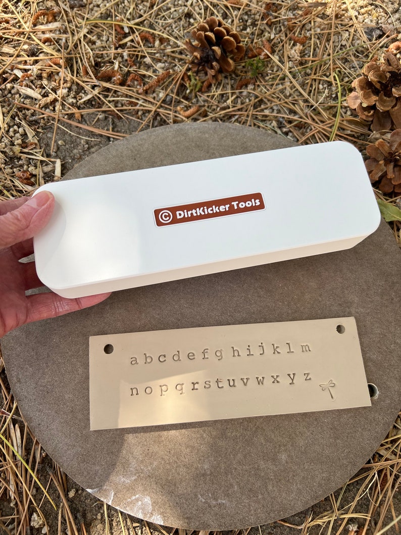 May include: A white rectangular box with the text "DirtKicker Tools" and a metal plate with stamped lowercase letters a-z and a dragonfly symbol. The items are on a gray stone.