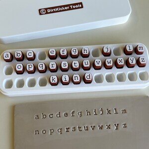 May include: A white case with the text "DirtKicker Tools" and a set of alphabet stamps. The stamps are brown with white letters, arranged in a white tray. A sample of stamped letters is also visible.