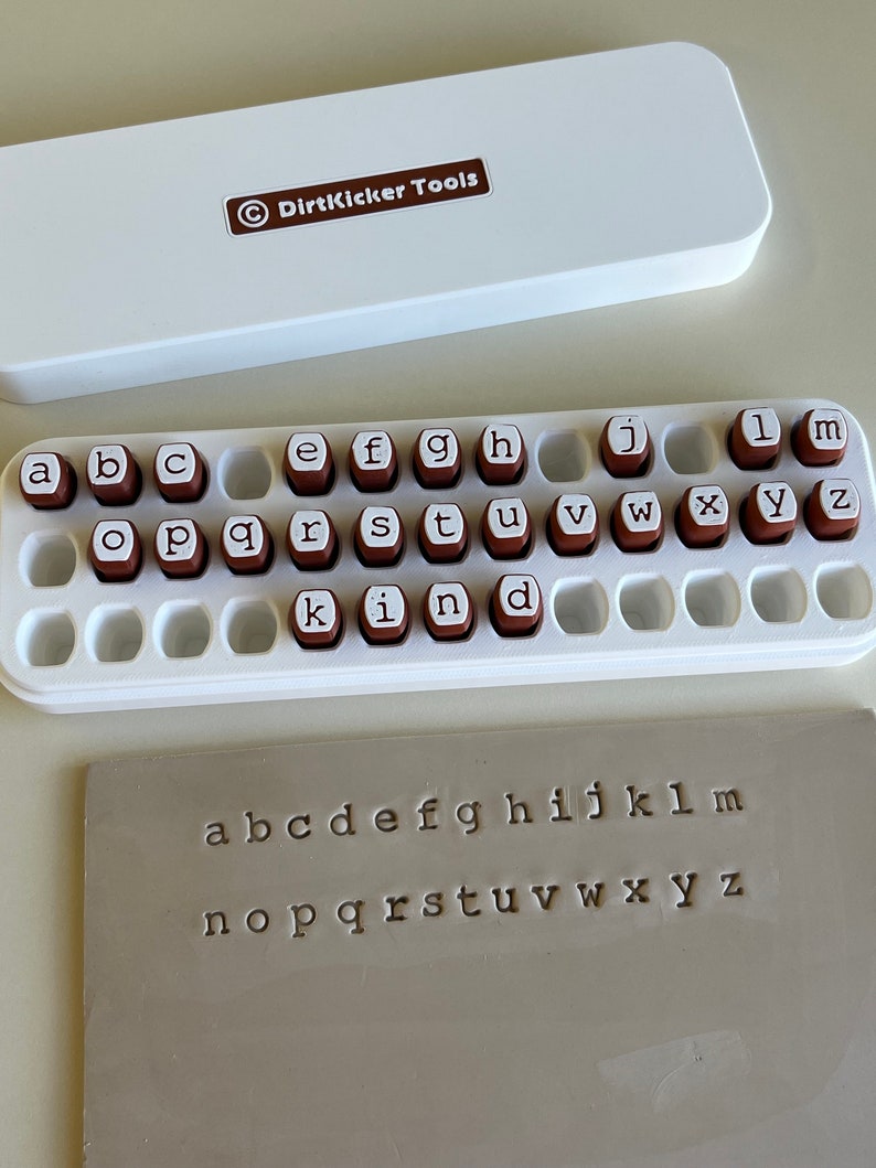 May include: A white case with the text "DirtKicker Tools" and a set of alphabet stamps. The stamps are brown with white letters, arranged in a white tray. A sample of stamped letters is also visible.