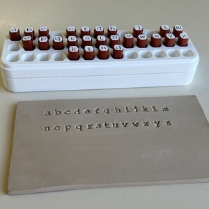 May include: A white plastic stamp holder with brown letter stamps and a gray rectangular stamp with the alphabet letters a through z stamped on it. The stamps are arranged in a white plastic holder.