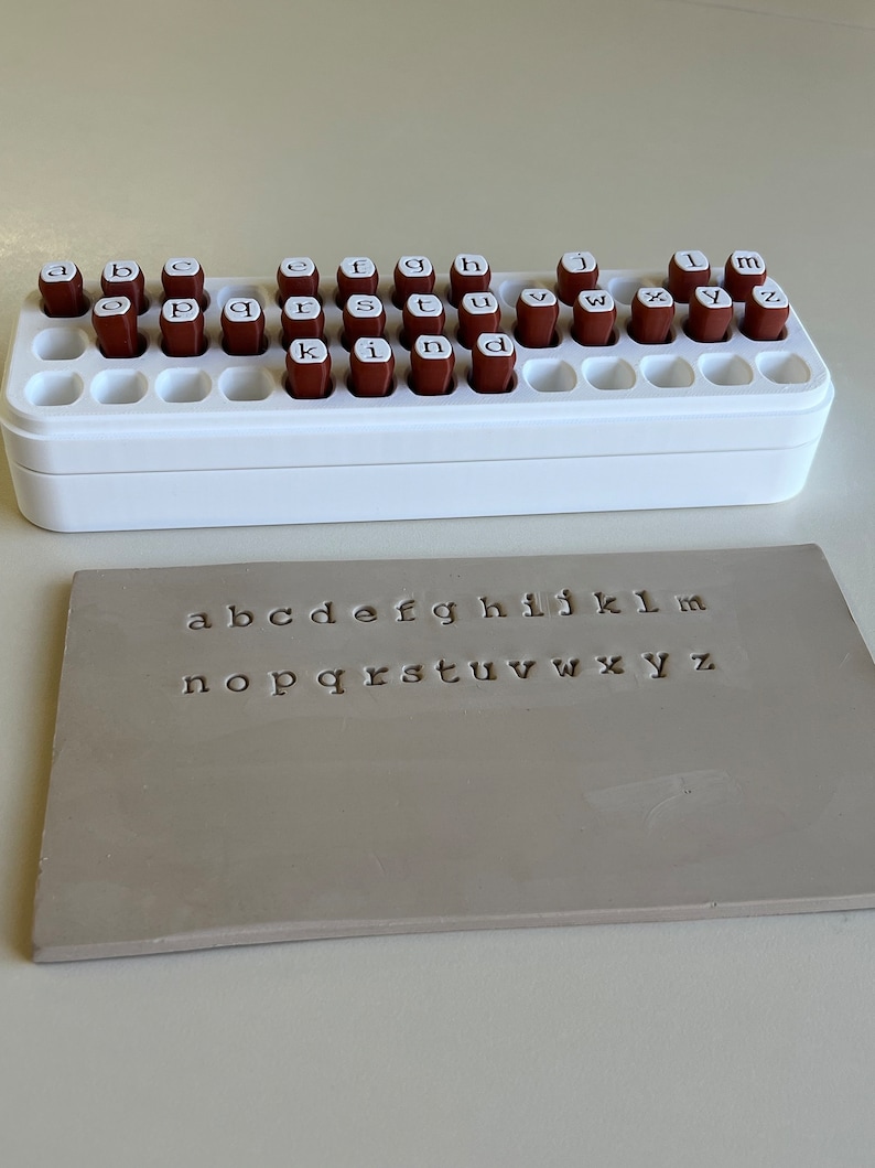 May include: A white plastic stamp holder with brown letter stamps and a gray rectangular stamp with the alphabet letters a through z stamped on it. The stamps are arranged in a white plastic holder.