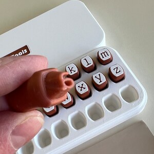 May include: A brown stamp handle is held above a white case containing individual letter stamps. The stamps are brown with white letter imprints. The case is open, revealing the stamps and a printed impression of the letters "k1m". The case has the word "Tools" printed on it.