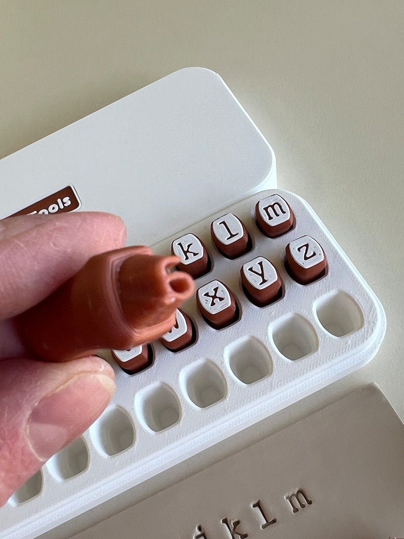 May include: A brown stamp handle is held above a white case containing individual letter stamps. The stamps are brown with white letter imprints. The case is open, revealing the stamps and a printed impression of the letters "k1m". The case has the word "Tools" printed on it.