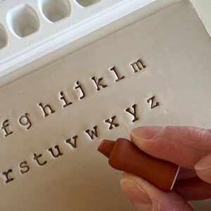 May include: A close-up of a letterpress printing set. The image shows a brown ink stamp being used to imprint the lowercase alphabet onto a beige surface. A white tray holds additional stamps.
