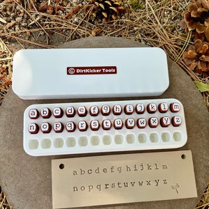 May include: A white rectangular case with the text "DirtKicker Tools" and a set of letter stamps. The stamps are red with white letters and are arranged in a white tray. A metal plate with stamped lowercase letters is also included.