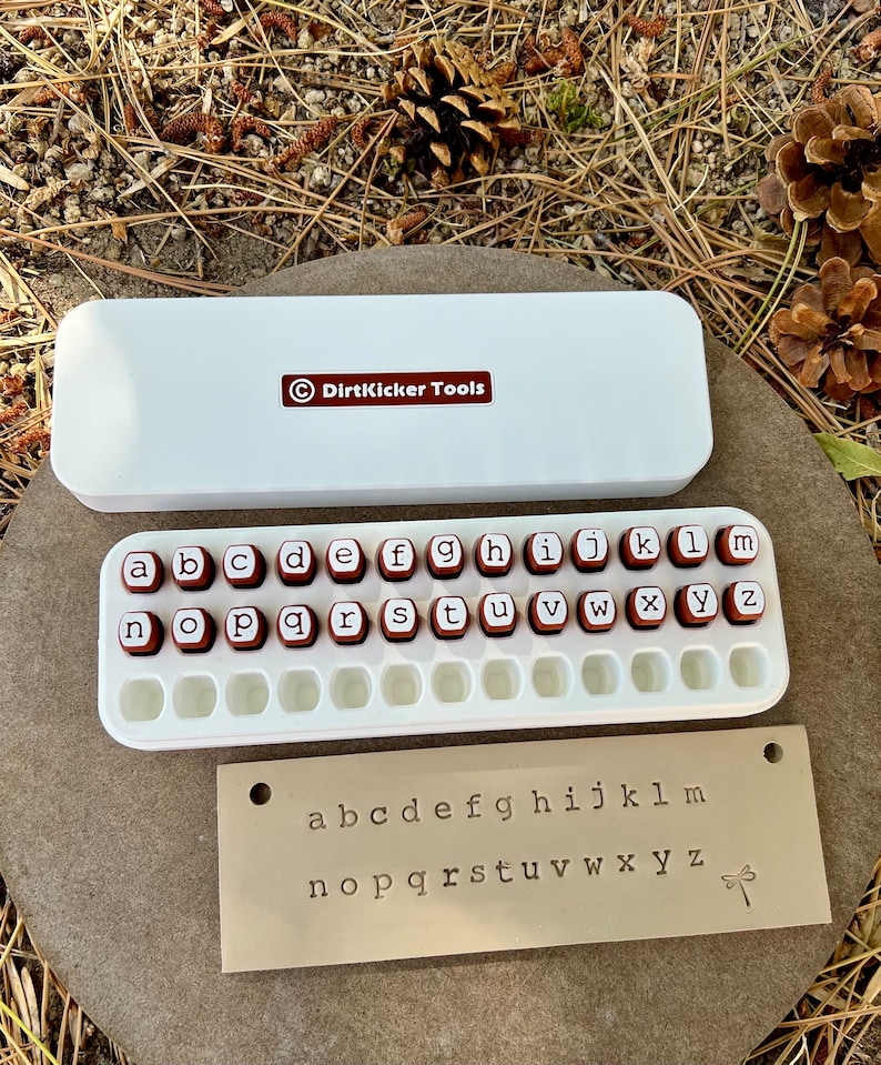 May include: A white rectangular case with the text "DirtKicker Tools" and a set of letter stamps. The stamps are red with white letters and are arranged in a white tray. A metal plate with stamped lowercase letters is also included.