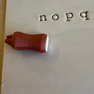 May include: A brown, cylindrical clay tool with a white tip rests on a gray surface with stamped lowercase letters. The tool appears to be a pottery shaping tool, used for detailed work.