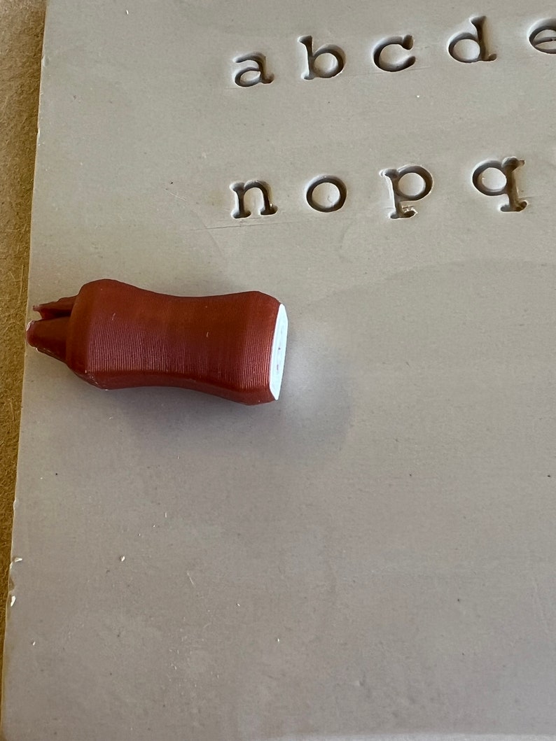 May include: A brown, cylindrical clay tool with a white tip rests on a gray surface with stamped lowercase letters. The tool appears to be a pottery shaping tool, used for detailed work.