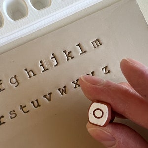 May include: A close-up of a letter stamp with the letter "O" on a light brown surface. The stamp is held by a person's hand. The background shows other letter stamps and the stamped letters "f g h i j k l m" and more.