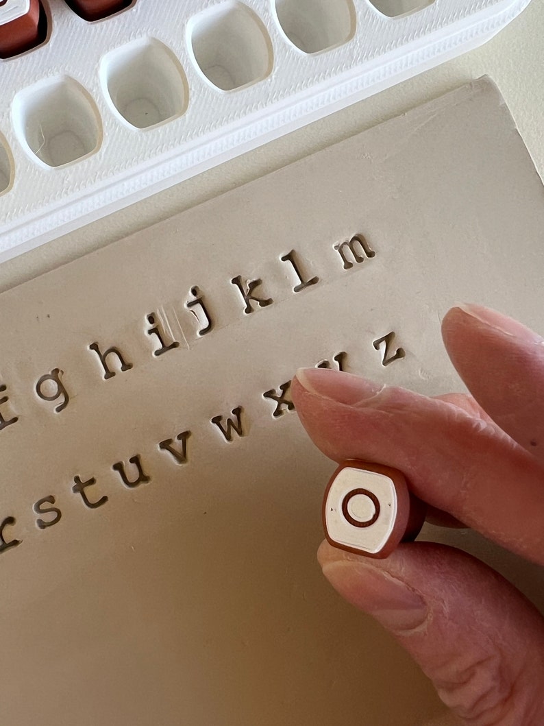 May include: A close-up of a letter stamp with the letter "O" on a light brown surface. The stamp is held by a person's hand. The background shows other letter stamps and the stamped letters "f g h i j k l m" and more.