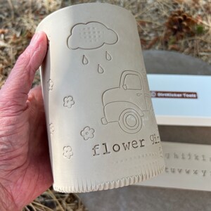 May include: A cylindrical, unglazed ceramic vase with embossed designs. The design includes a cloud with raindrops, a vintage truck, and the words "flower girl." Small flower motifs are scattered around the vase. The vase is held in a hand.