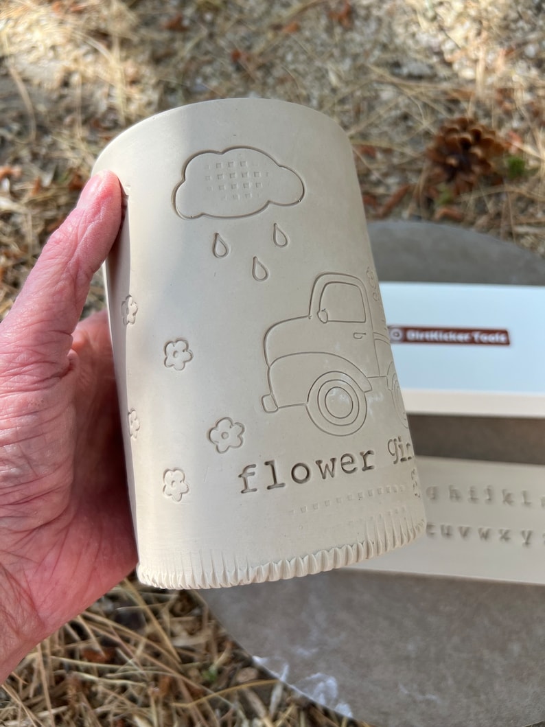 May include: A cylindrical, unglazed ceramic vase with embossed designs. The design includes a cloud with raindrops, a vintage truck, and the words "flower girl." Small flower motifs are scattered around the vase. The vase is held in a hand.