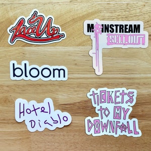 Op de afbeelding: Vijf stickers op een houten oppervlak. De stickers zijn wit met zwarte en roze letters. De stickers zeggen "Face Up", "Bloom", "Hotel Disco", "Mainstream Sell Out" en "Tickets to My Downfall".
