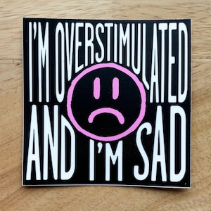 Op de afbeelding: Een vierkante sticker met de tekst "I'M OVERSTIMULATED AND I'M SAD" in het wit, met een roze verdrietig gezicht in het midden. De achtergrond is zwart.