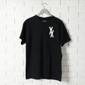May include: Black t-shirt with a white "XX" graphic on the front. The shirt is hanging on a hanger against a white brick wall.
