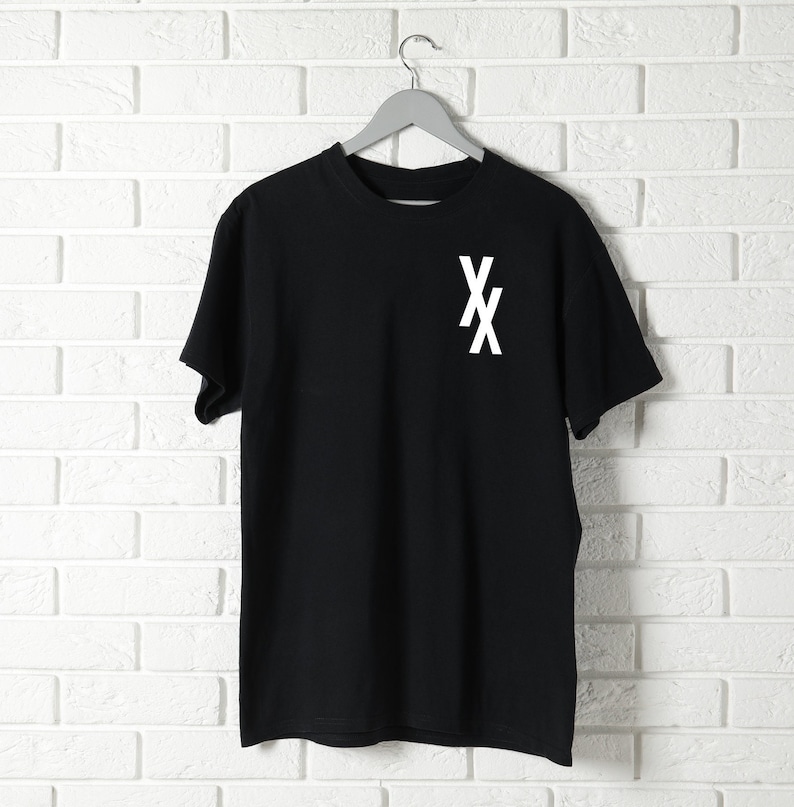 May include: Black t-shirt with a white "XX" graphic on the front. The shirt is hanging on a hanger against a white brick wall.