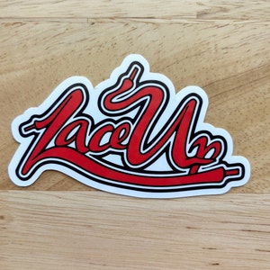 Op de afbeelding: Rode en zwarte sticker met het woord "Lace Up" geschreven in een gestileerd lettertype. Het woord wordt gevormd door een rode veters.