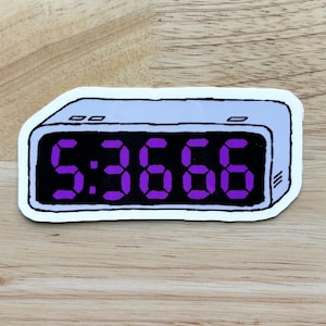 Può includere: Un orologio digitale bianco con un quadrante nero e numeri viola che mostrano l'ora 5:3668.