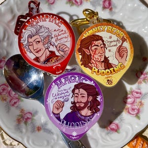 Puede incluir: Tres colgantes acrílicos coloridos con ilustraciones de personajes, cada uno etiquetado como "Vampire Pudding", "Honeybear Pudding" y "Magic Pudding". Los colgantes están sobre un plato decorativo con una cuchara, rodeados de pequeños alimentos decorativos.