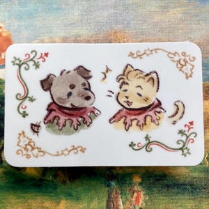 May include: A rectangular sticker featuring a watercolor illustration of a dog and a cat. The dog is gray, and the cat is tan, both wearing red ruffled collars. Decorative vines and berries frame the image on a white background.