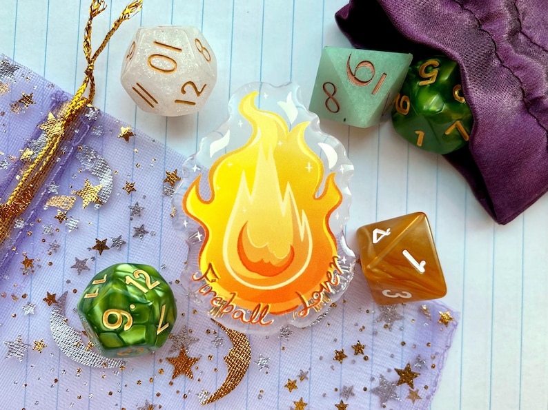 May include: A set of dice with a fireball design. The dice are green, white, and brown with gold numbers. The fireball design is on a clear acrylic background with the words "Fireball" and "Lover" written in gold.