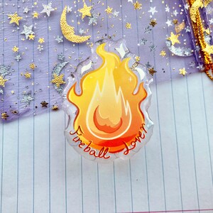 May include: A clear, round, resin charm with a yellow and orange flame design. The charm has the words "Fireball Lover" written around the edge.