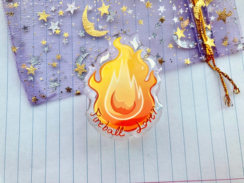 May include: A clear, round, resin charm with a yellow and orange flame design. The charm has the words "Fireball Lover" written around the edge.