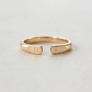 May include: A gold-colored ring with an open design. The ring features the letters "B" and "P" engraved on the ends. The ring is set against a neutral background, highlighting its simple elegance.