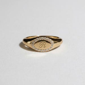Könnte beinhalten: Ein goldener Siegelring mit ovaler Oberfläche. Die Oberfläche ist von kleinen, klaren Steinen umgeben und zeigt den kursiven Buchstaben "R". Der Ring ist vor einem weißen Hintergrund platziert.
