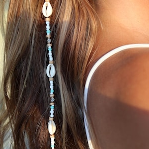 May include: A white and brown beaded hair tie with small white seashells. The hair tie is worn in a woman's brown hair.