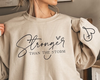May include: A beige sweatshirt with the text "Stronger than the storm" and a small heart design. The right sleeve has a small anchor design.
