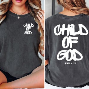 May include: Dark gray t-shirt with the words "CHILD OF GOD" printed in white on the front and back. The back of the shirt also includes "PSALM 23" in white. The shirt has a vintage, worn look.