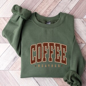 Puede incluir: Una sudadera verde con el texto "COFFEE WEATHER" en letras marrones.