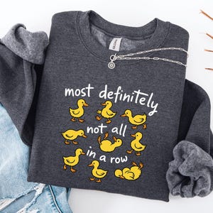 May include: A dark gray sweatshirt with the phrase "most definitely not all in a row" and cartoon yellow ducklings. A silver necklace with a circular pendant is draped over the sweatshirt. The sweatshirt is folded on top of a pair of blue jeans.
