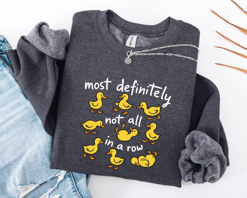 May include: A dark gray sweatshirt with the phrase "most definitely not all in a row" and cartoon yellow ducklings. A silver necklace with a circular pendant is draped over the sweatshirt. The sweatshirt is folded on top of a pair of blue jeans.
