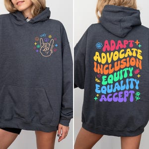 May include: A dark gray hoodie with a rainbow graphic on the front and the words "Adapt, Advocate, Inclusion, Equity, Equality, Accept" on the back.