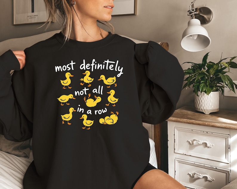 May include: Black sweatshirt with the phrase "most definitely not all in a row" in white text. The design features yellow cartoon ducklings. A comfortable, casual top.