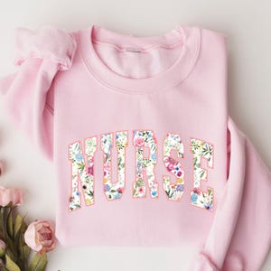May include: Pink sweatshirt with the word "NURSE" in floral print letters.