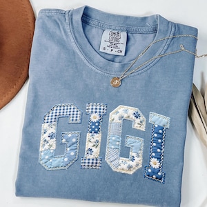 May include: A light blue Comfort Colors t-shirt with the name "GIGI" appliqued in patchwork fabric. The letters feature floral, gingham, and polka dot patterns in shades of blue and white. A gold necklace with a pendant rests on the shirt.