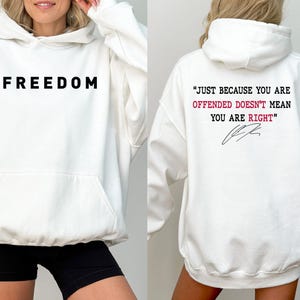 May include: White hoodie with the word "FREEDOM" printed in black on the front. The back of the hoodie features the quote, "JUST BECAUSE YOU ARE OFFENDED DOESN'T MEAN YOU ARE RIGHT" in red and black text, with a signature.